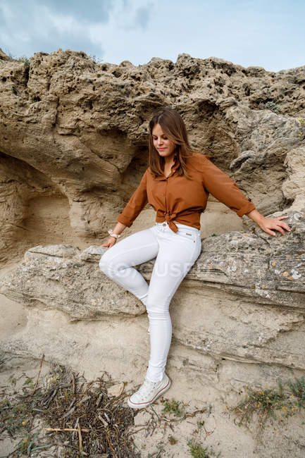 Hermosa mujer aventurera de vaqueros blancos sentada sobre piedras rocosas en el desierto. - foto de stock