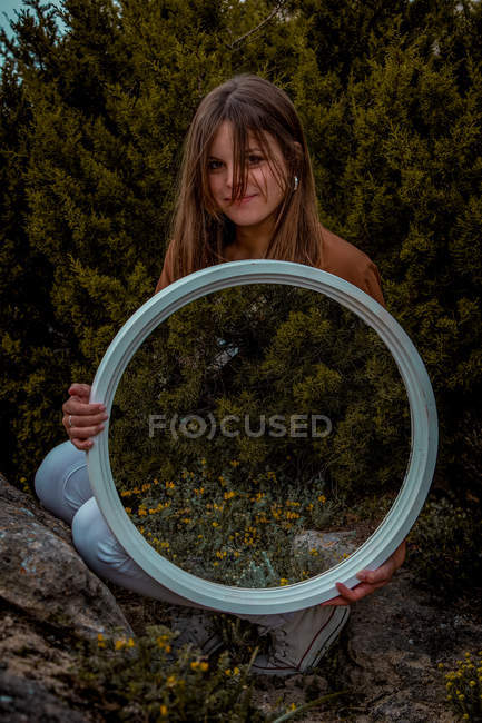 Schöne Frau steht mit großem runden Spiegel in der Nähe grüner Büsche und blickt in die Kamera — Stockfoto