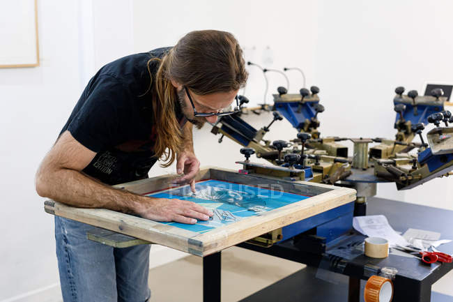 Artista masculino concentrado em avental sujo trabalhando com serigrafia enquanto cria estampa em t-shirts em oficina — Fotografia de Stock