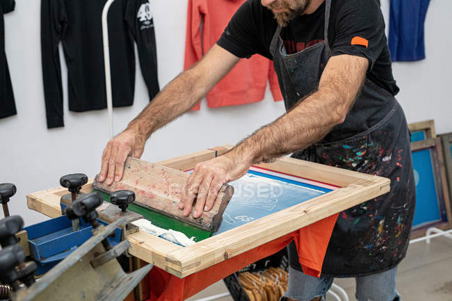 Erntehelfer in schmutziger Schürze arbeitet mit Siebdruck, während er in der Werkstatt Druck auf T-Shirt erzeugt — Stockfoto