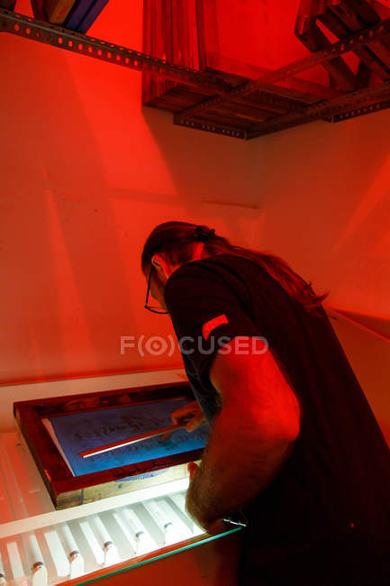 Focused male artist with long hair drying printing screen red illuminated room of workshop — Stock Photo
