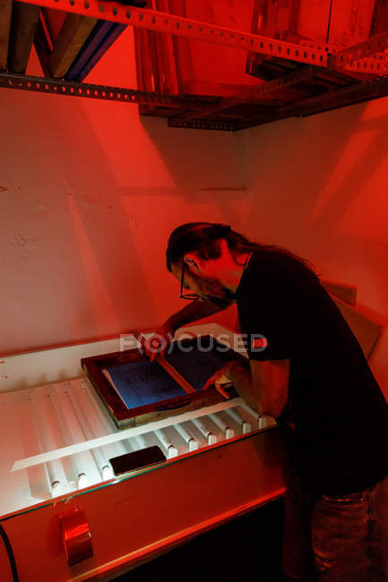 Focused male artist with long hair drying printing screen red illuminated room of workshop — Stock Photo