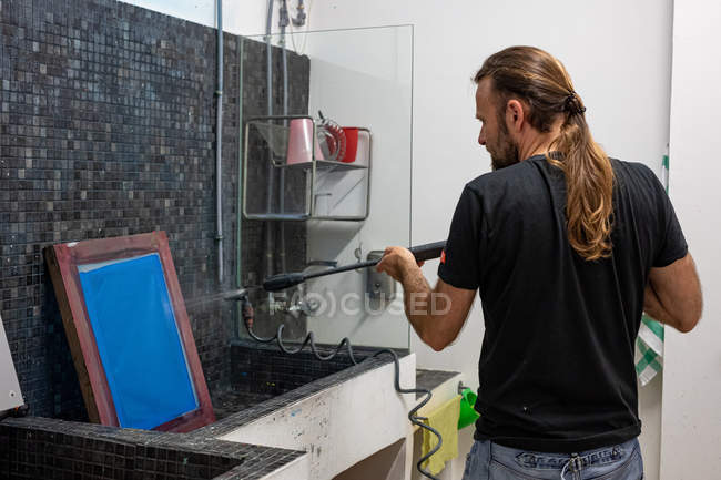 Окаменевший художник-мужчина с длинными волосами готовит синий шелкограф для печати на экране в мастерской — стоковое фото