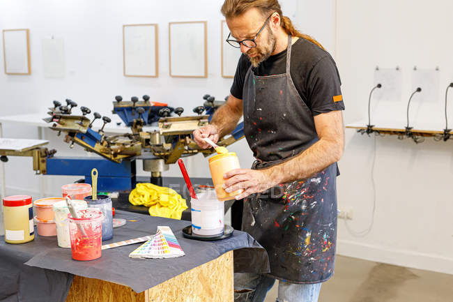 Concentrated man with long hair and glasses wearing dirty apron mixing various paints for serigraphy in workshop — Stock Photo