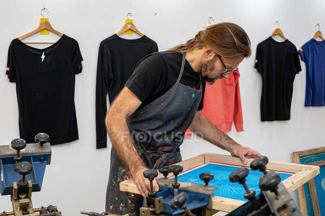 Концентрований чоловічий художник у брудному фартусі, що працює з шовкографією, створюючи друк на футболках у майстерні — стокове фото
