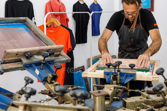 Концентрированный художник-мужчина в грязном фартуке работает с шелкографией, создавая принты на футболках в мастерской — стоковое фото