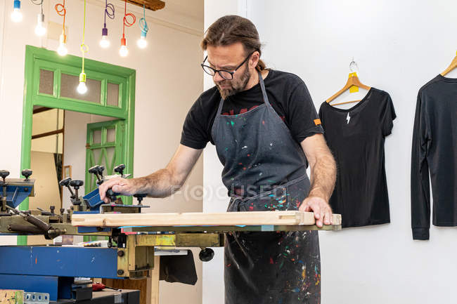 Artista masculino concentrado en el delantal sucio trabajando con pantalla de seda mientras se crea impresión en camisetas en taller. - foto de stock