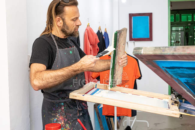 Konzentrierter männlicher Künstler in schmutziger Schürze arbeitet mit Siebdruck, während er in der Werkstatt T-Shirts bedruckt — Stockfoto