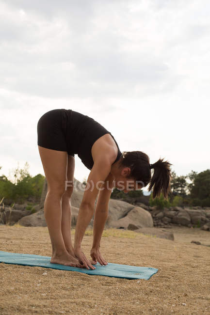 Mulher adulta média se curvando para a frente enquanto faz ioga ao ar livre na praia da barragem — Fotografia de Stock