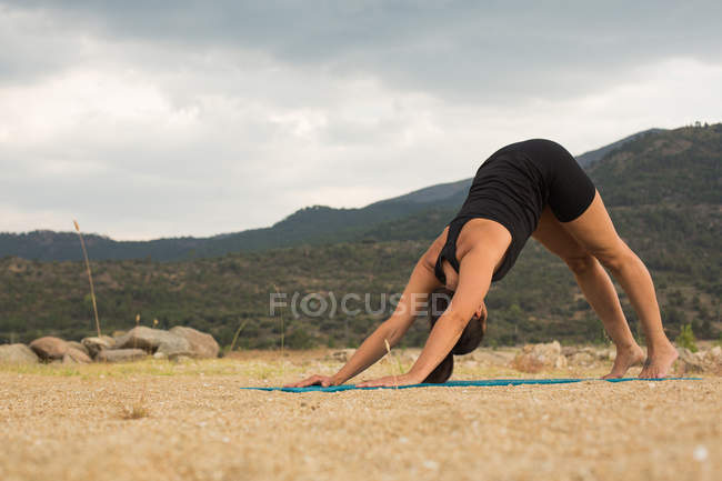 Femme adulte moyenne en posture de chien orientée vers le bas tout en faisant du yoga à l'extérieur sur la plage du barrage — Photo de stock