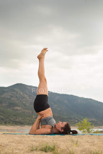 Mulher adulta média no suporte de ombros enquanto faz ioga ao ar livre na praia da barragem — Fotografia de Stock