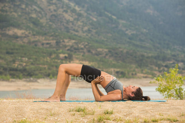 Середня доросла жінка в позі мосту робить йогу на відкритому повітрі на пляжі греблі — стокове фото
