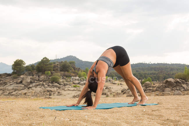 Mid adult woman in downward-facing dog pose while doing yoga outdoors on dam beach — Stock Photo