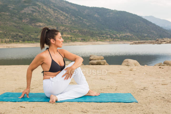 Mujer adulta media estirándose mientras hace yoga al aire libre en la playa de la presa - foto de stock