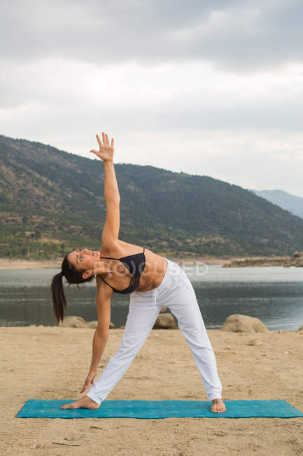 Mid adult woman in triangle pose doing yoga outdoors on dam beach — Stock Photo