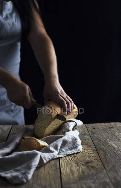 Unrecognizable female cutting loaf of fresh bread on napkin while standing near old wooden table on black background — Stock Photo