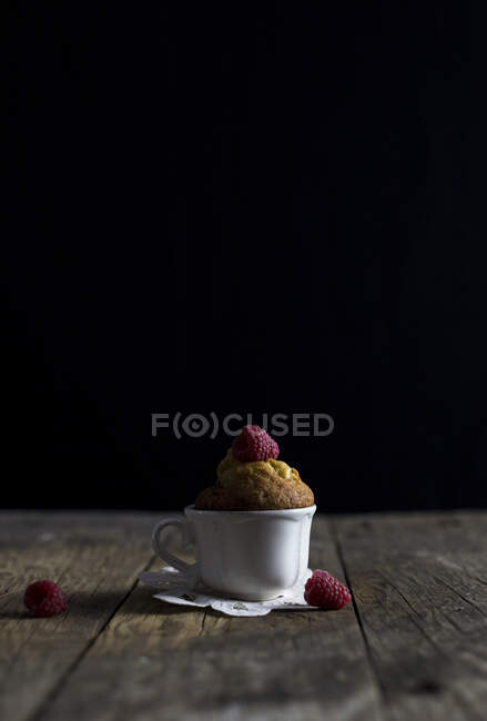 Ceramic cup with yummy raspberry muffin placed on shabby timber tabletop against black background — Stock Photo