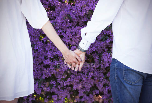 Анонимные мужчина и женщина держатся за руки, стоя возле кустарника с фиолетовыми цветами в парке — стоковое фото