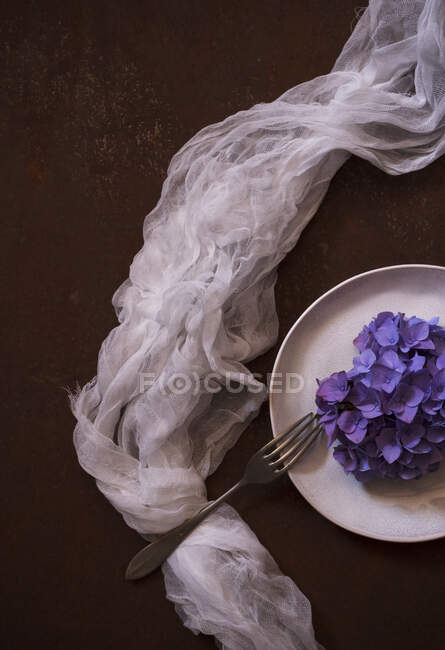 Зверху тонка напівпрозора тканина, розміщена біля тарілки з купою фіолетових квітів на коричневій поверхні — стокове фото