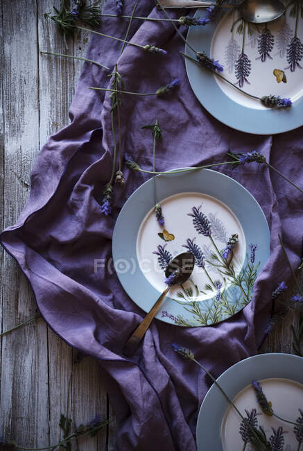 From above fresh lavenders placed around empty plates over violet fabric on lumber tabletop — Stock Photo