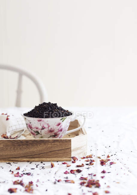 Porzellantasse voller getrockneter Teeblätter, die morgens auf einem Holztablett in der Nähe von Blütenblättern auf dem Tisch stehen — Stockfoto