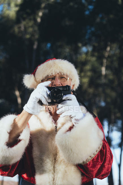 Smiling senior man in costume of Santa Claus standing and taking photos with camera on nature background — Stock Photo
