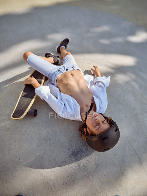 Vue d'en haut de l'enfant dans un casque couché les yeux fermés et se refroidissant au sol dans un skatepark aux ombres — Photo de stock