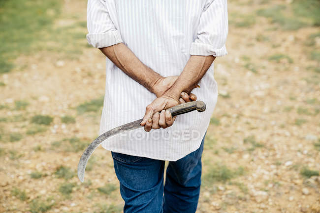 Casual man carrying sickle while walking along dry field in daylight — Stock Photo
