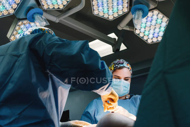 D'en bas jeune médecin sérieux dans le masque de protection et la casquette faisant la chirurgie avec des instruments et infirmière de culture — Photo de stock