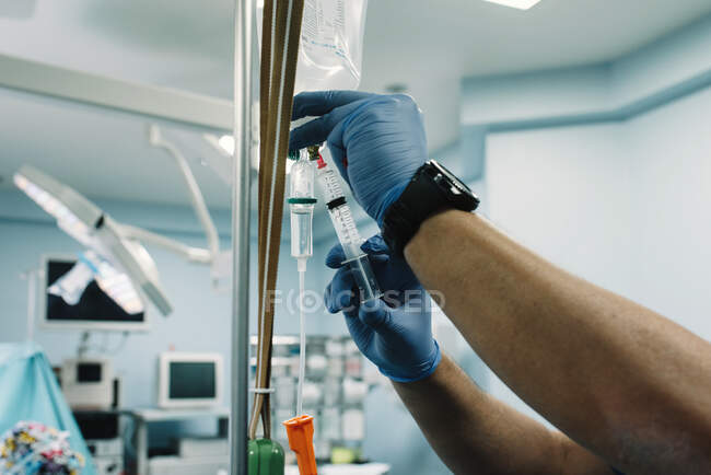 Crop hands in rubber gloves adding medication in saline solution drip with sterile syringe in hospital — Stock Photo