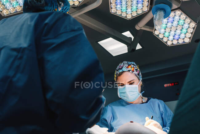 Von unten ernsthafter junger Arzt in Schutzmaske und Mütze, der mit Instrumenten und Erntekrankenschwester operiert — Stockfoto