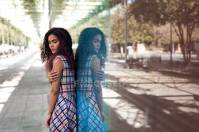 Side view of young ethnic woman embracing herself standing near glass wall with reflection on street — Stock Photo