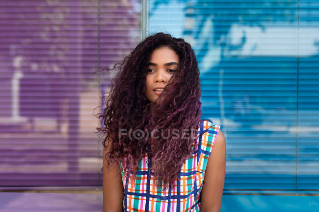 Sensuelle jeune femme ethnique en robe colorée appuyée sur le mur de verre coloré bleu et rose détournant les yeux — Photo de stock