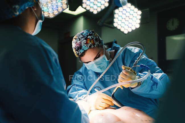 Sérieux jeune médecin en masque protecteur et casquette faisant la chirurgie avec des instruments et infirmière de culture — Photo de stock