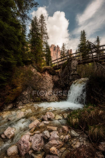 Fundo rochoso com corrente de água fluindo sob ponte de madeira através de floresta sombria em montanhas em dia nublado em Dolomites, Itália — Fotografia de Stock