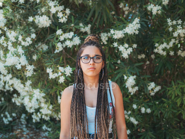 Молода жінка в окулярах і літньому одязі стоїть біля квітучих дерев і дивиться на камеру — стокове фото
