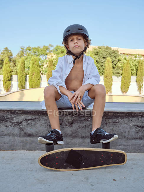 Niño casual con casco y camisa blanca sentado en la rampa en skatepark mirando en la cámara - foto de stock