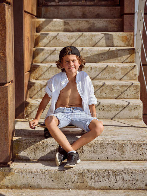 Menino alegre sentado no skate nas escadas no skatepark no verão ensolarado durante o dia olhando na câmera — Fotografia de Stock