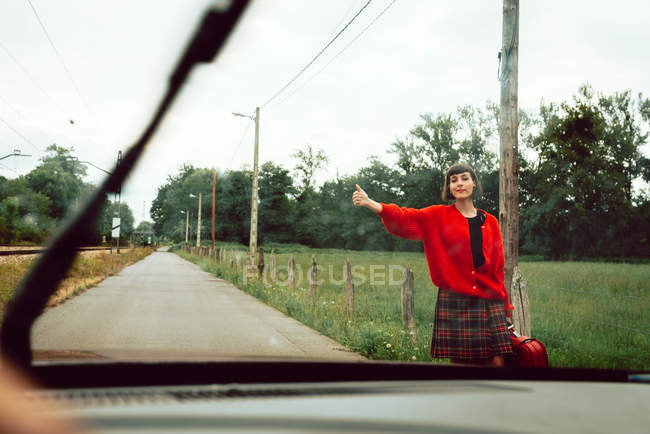 Femme en pull rouge attrapant voiture sur la route dans la campagne — Photo de stock