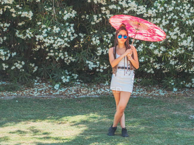 Schlanke junge Frau im Sommer-Outfit mit Regenschirm, die Beine in der Nähe blühender Bäume gekreuzt — Stockfoto