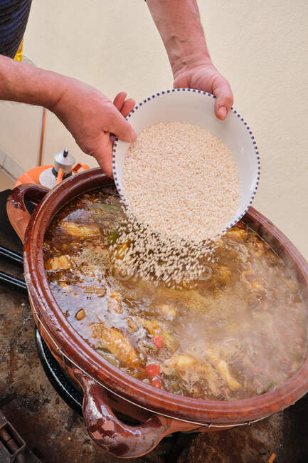 Cortar las manos de un humano anónimo vertiendo arroz en una olla grande de barro con un plato caliente de verduras de pollo y caracoles de Borgoña - foto de stock