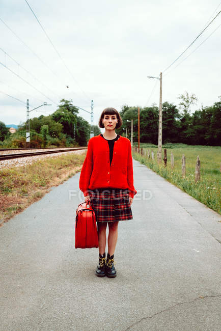 Жінка в стильних чоботях з червоною валізою, що стоїть на дорозі в сільській місцевості — стокове фото