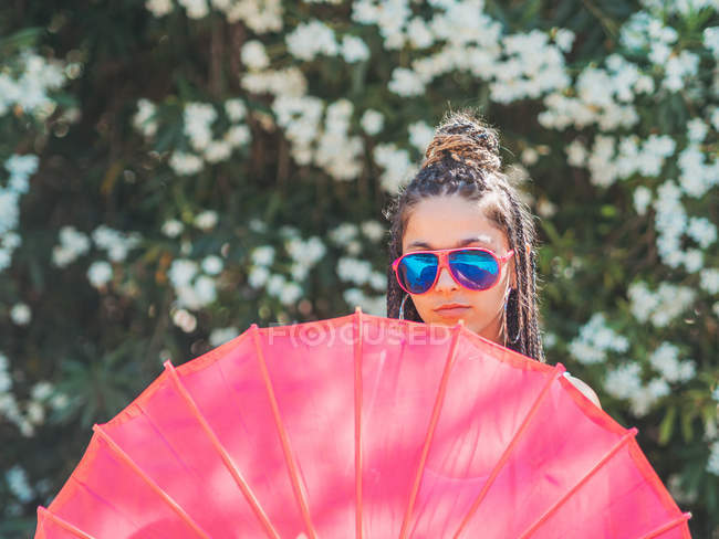 Slim young woman in sunglasses with umbrella standing near blooming trees — Stock Photo