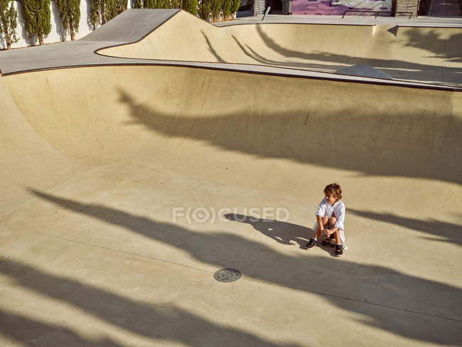 Vue d'en haut de l'enfant dans le casque assis et se refroidissant sur le sol dans le skatepark avec des ombres regardant loin — Photo de stock