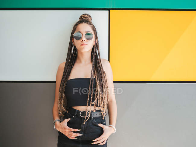Pretty stylish teenage girl with hands in pockets and unique dreadlocks looking at camera on colorful background — Stock Photo