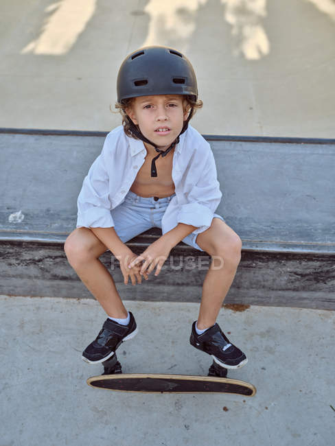 Freizeitkind mit Helm und weißem Hemd sitzt auf Rampe im Skatepark und schaut in die Kamera — Stockfoto