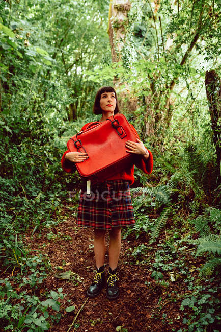Woman in red with vintage red suitcase standing in forest — Stock Photo