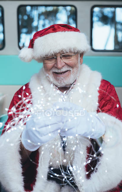 Старший в костюме Санта-Клауса сидит в ретро-фургоне и держит гирлянду в руках в перчатках — стоковое фото