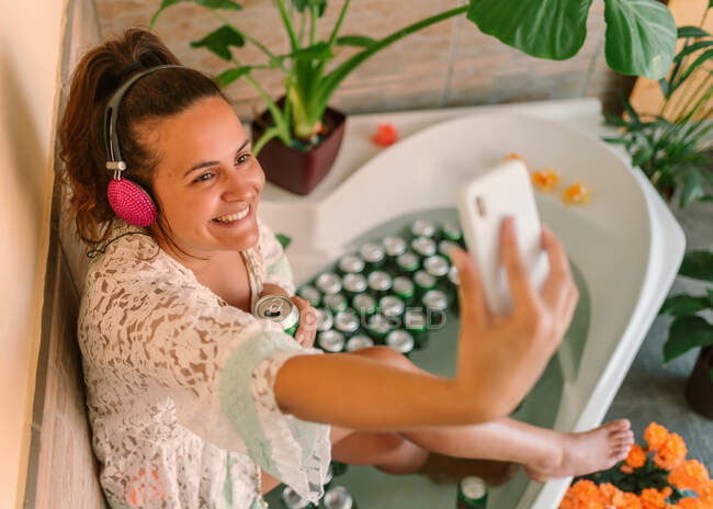 Вид сверху на жизнерадостную молодую женщину в наушниках с пивной банкой в ванной, говорящую селфи на смартфоне — стоковое фото