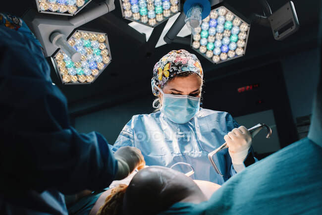 Знизу серйозного молодого лікаря в захисній масці і капелюсі роблять операцію з інструментами і медсестрою врожаю — стокове фото